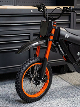 Load image into Gallery viewer, Close-up of a black and orange electric dirt bike or motorcycle front wheel, fender, handlebar area, and battery compartment, photographed in a workshop setting with a gray tool chest behind it.