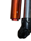 Close-up of a metal mechanical component with an anodized orange-red cylindrical section and a black plastic or cast-metal joint, placed on a wood surface.