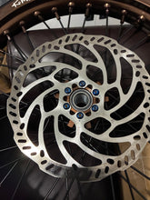 Load image into Gallery viewer, A close-up of a metallic silver disc brake rotor mounted on a bicycle or motorcycle wheel hub with visible spokes and blue lock nuts.
