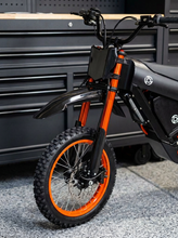 Load image into Gallery viewer, A close-up of a black and orange electric dirt bike or moped front wheel, showing the knobby tire, disc brake, fender, battery, and handlebars, positioned in front of a dark gray tool chest.

