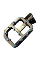 Load image into Gallery viewer, A black metal bicycle or motorcycle crank arm with a matching flat platform pedal featuring blue anodized accents and blue-tipped bolts, resting on a dark gray textured surface.
